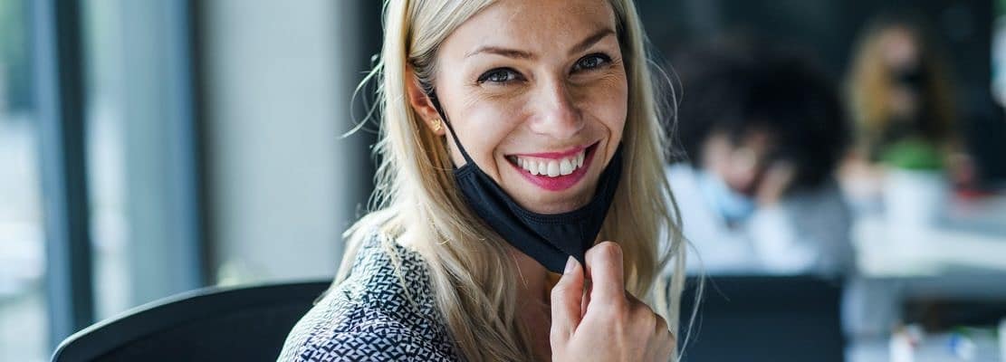 Young woman with face mask back at work in office after lockdown, looking at camera