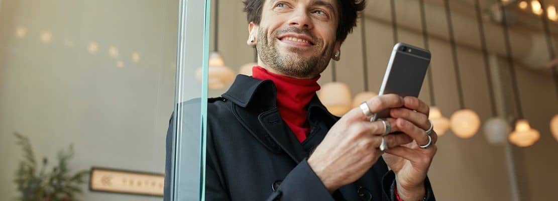 Pleased young handsome brown haired with earpieces keeping smartphone and looking cheerfully