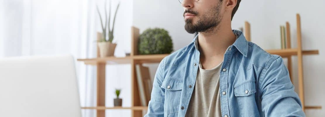 Thoughtful young copywriter being deep in thoughts, sits in front of opened laptop, thinks on creati