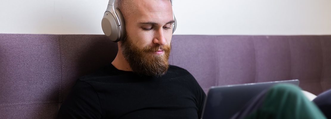 Caucasian bearded man in headphones in bedroom on bed working on laptop from home, typing, thinking.