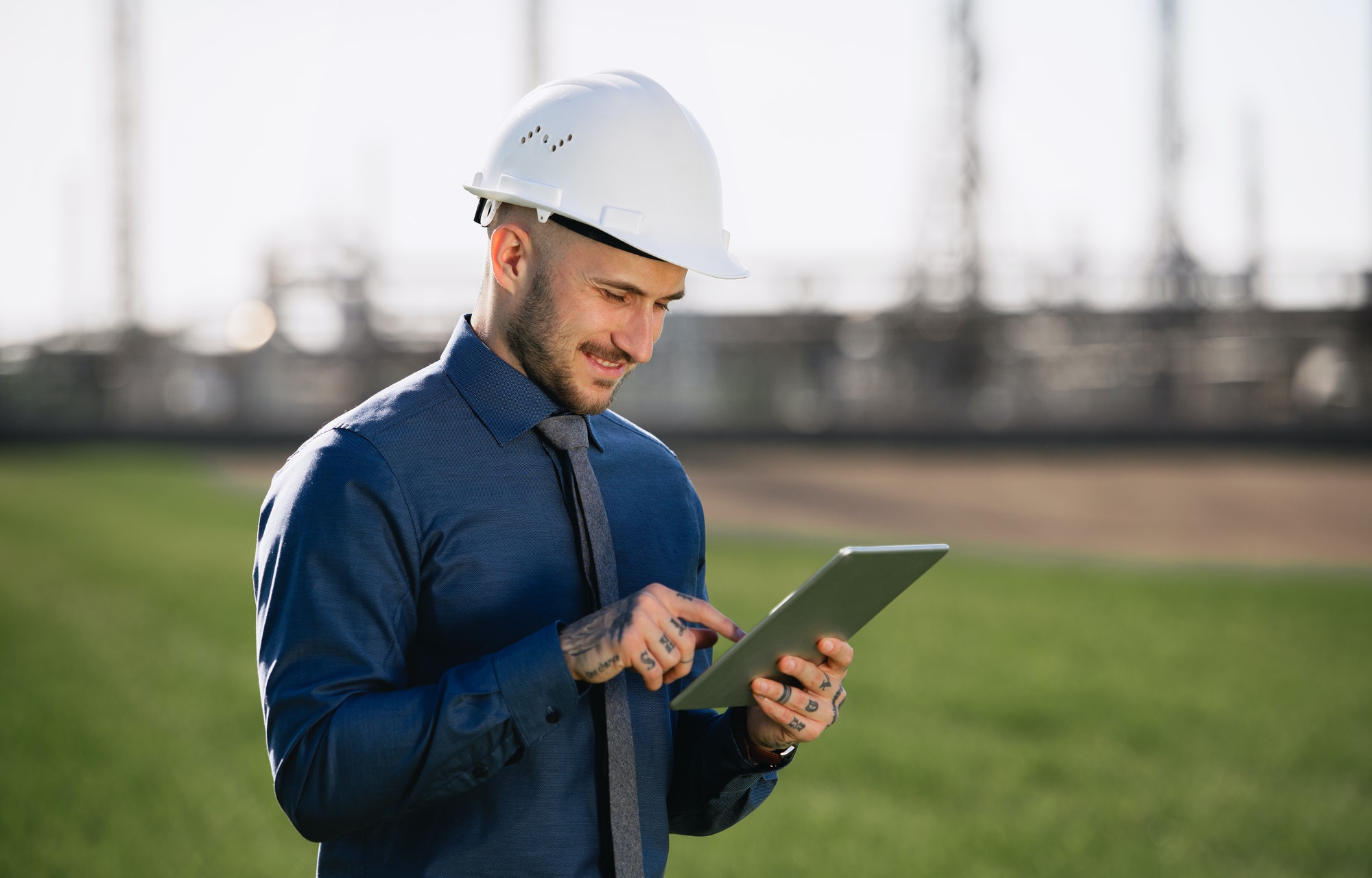 Young engineer with hard hat and tablet standing outdoors by oil refinery