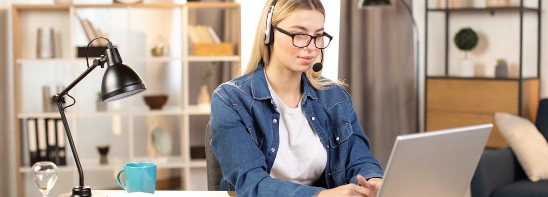 Pretty young woman using headset and laptop for video call, working at home
