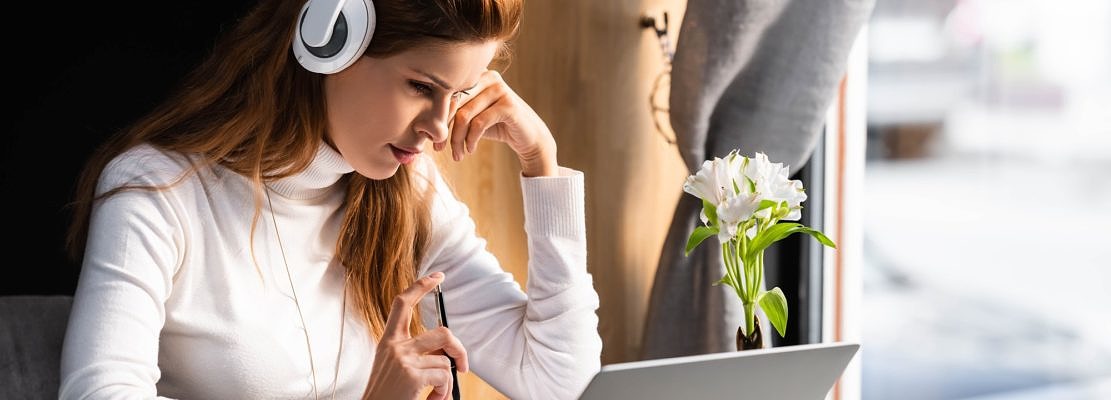 beautiful thoughtful woman in headphones watching webinar on laptop in cafe
