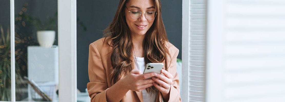 Young business woman in glasses with long hair in beige suit using smartphone office