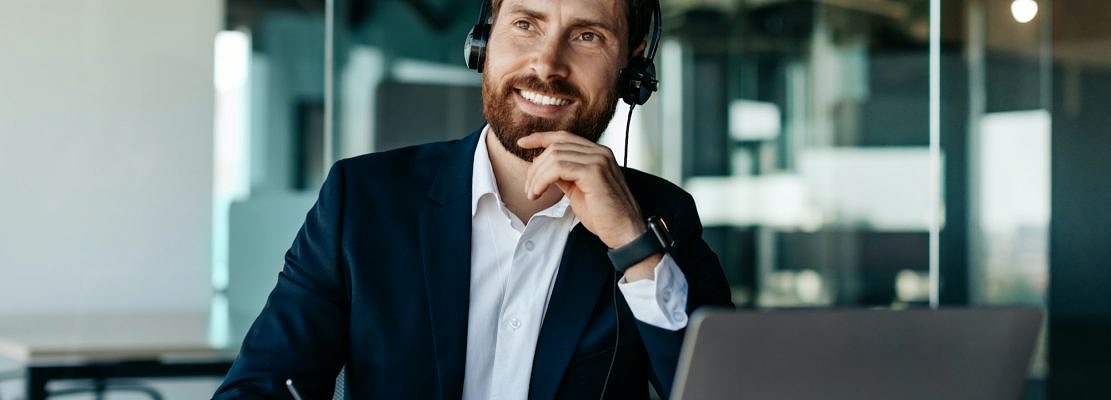 Happy thoughtful businessman in headset working on laptop and writing in notebook, sitting at desk