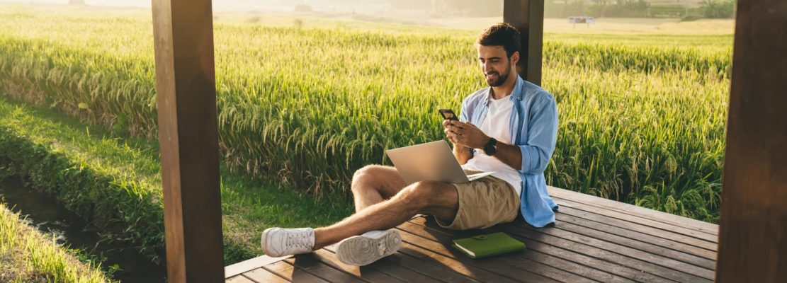Cheerful young man working distantly in countryside using laptop and smartphone