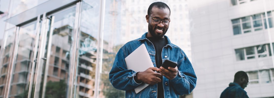 Cheerful hipster guy with netbook using mobile app for checking internet connection on cellphone