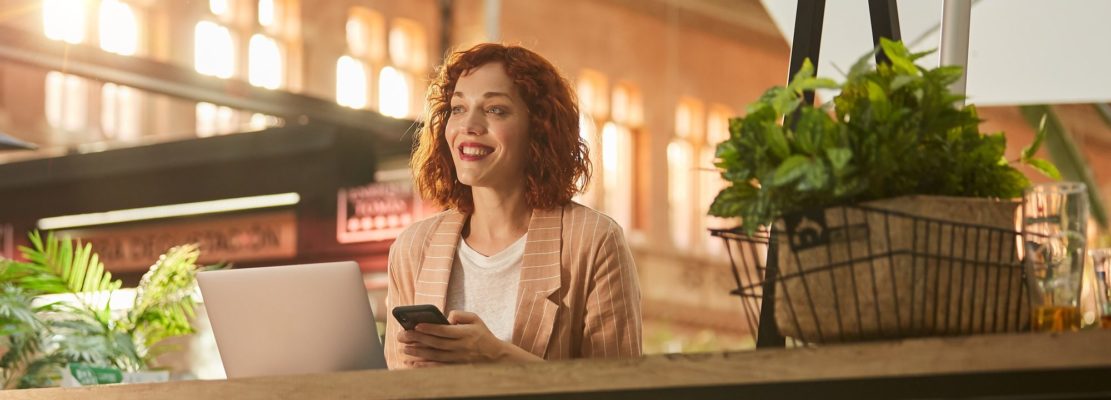 Cheerful young woman working on laptop in cafe and browsing phone