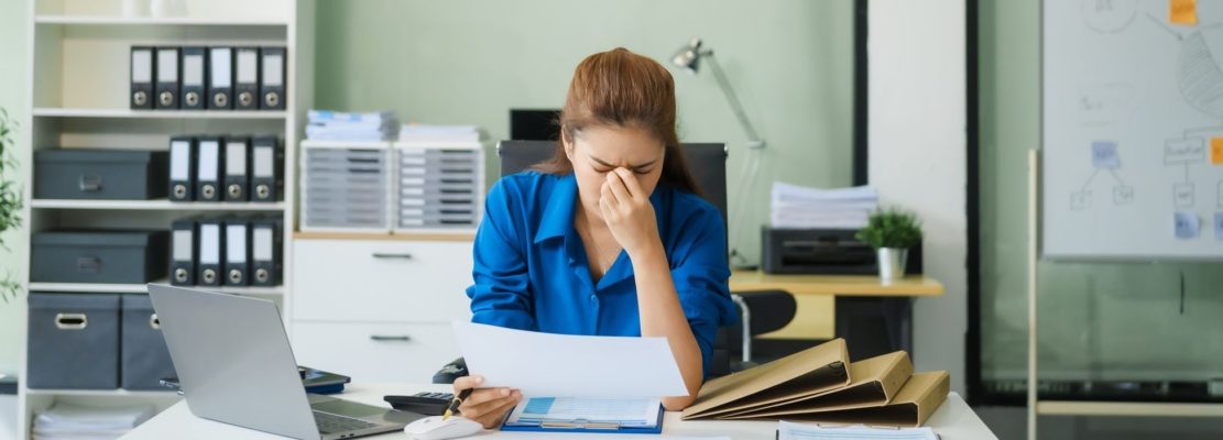 A businesswoman working at a desk in an office struggles with stress headaches caused by Office