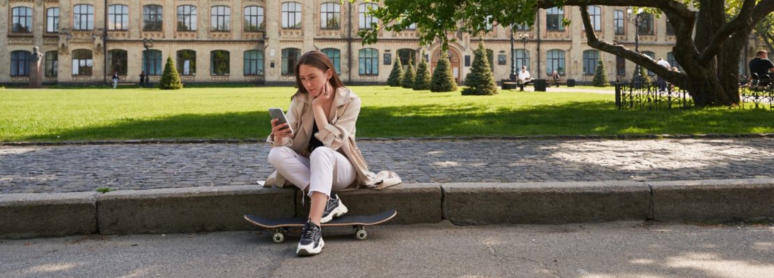 Lady sits on sidewalk in park and uses a smartphone