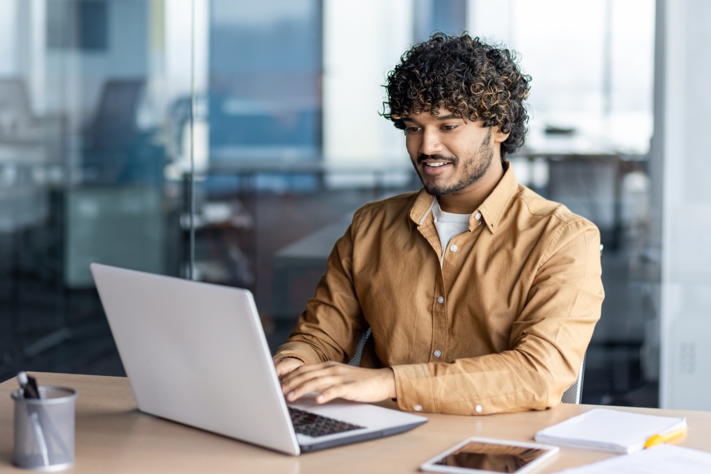 Upskilling und Reskilling: Wie eLearning die globale Qualifikationslücke schließen kann 2 Happy and smiling hispanic businessman typing on laptop, office worker with curly hair happy with
