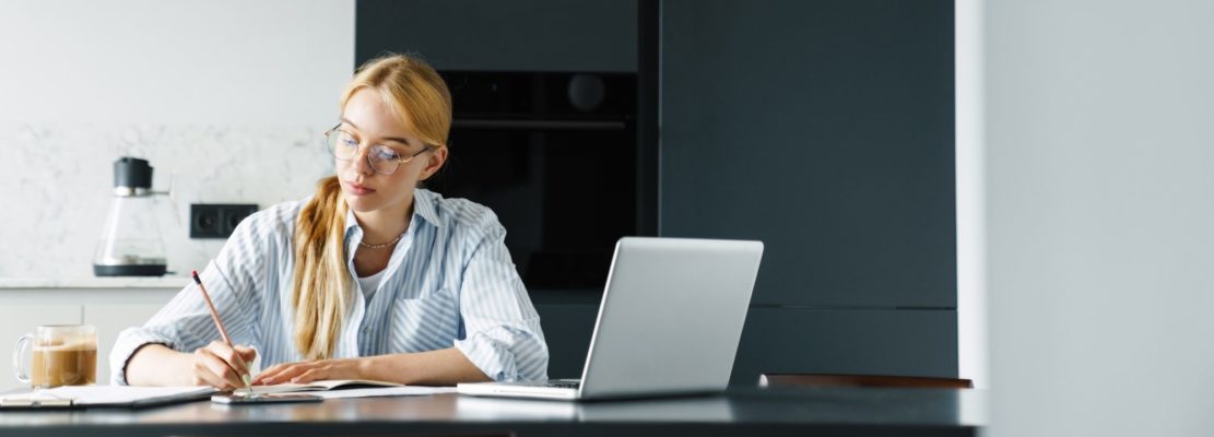 Photo of focused woman making down notes while working with laptop