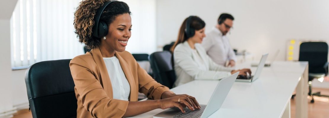 Beautiful African businesswoman wearing headphones working on laptop in co-working office, portrait.