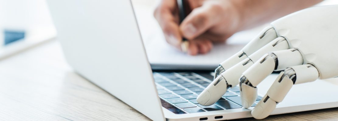 close-up view of robotic arm using laptop and human hand taking notes at wooden table