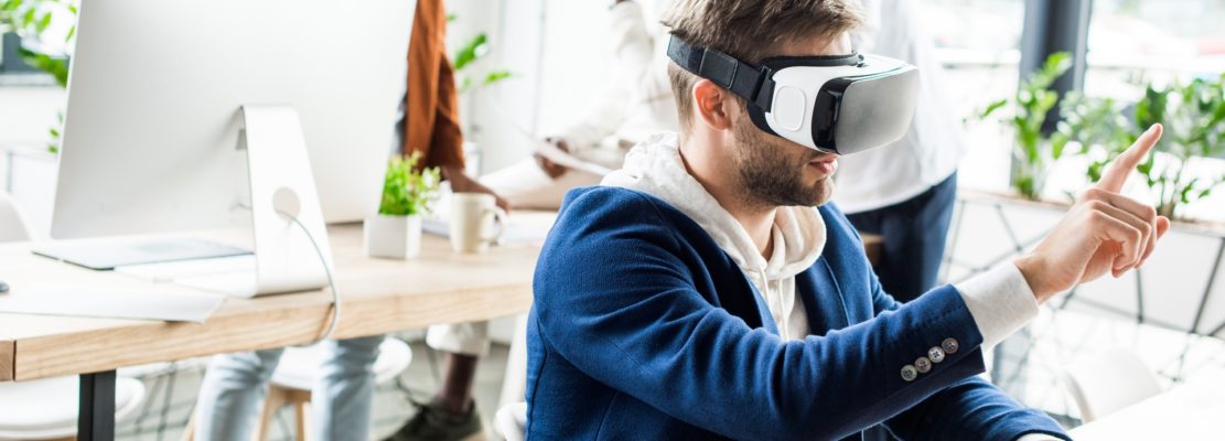 panoramic shot of young businessman touching something with finger while using vr headset in office